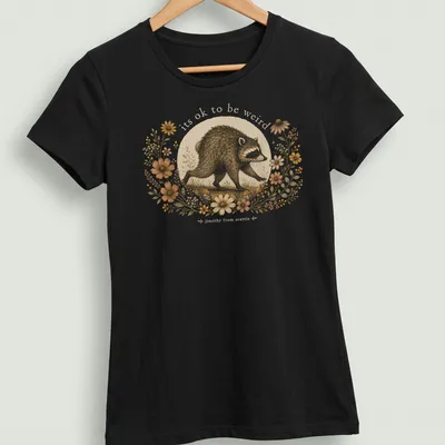 Cottagecore style T-shirt with a whimsical raccoon and the text 'its ok to be weird jimothy from seattle' — printed on a women's shirt